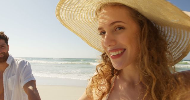 A young Caucasian woman wearing a sunhat smiles brightly on a sunny beach day, with a young Caucasian man partially visible in the background, with copy space. Their relaxed attire and the seaside setting suggest a leisurely vacation or a summer getaway.