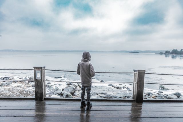 Person stands alone on a wooden pier, staring out at an icy lake surrounded by a winter landscape. The cloudy weather and foggy sky create a calm and reflective atmosphere, conveying themes of solitude and tranquility. Ideal for use in articles, blogs, and personal narratives about solitude, winter, cold weather, mental calmness, or scenic landscape photography.