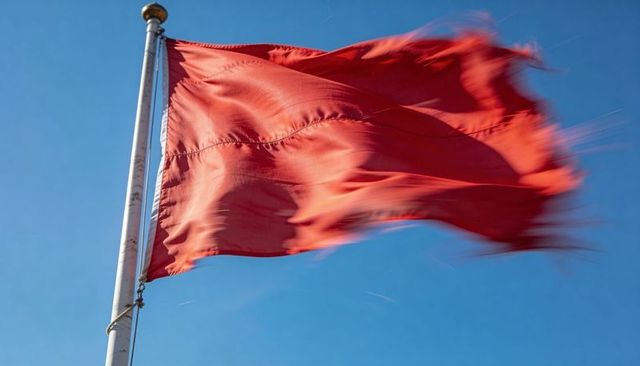 Waving crimson flag on metal pole against clear blue sky, wind motion and fabric texture