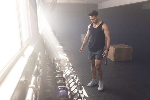 Male toned athlete inspecting dumbbell rack in sunlit gym wearing smartwatch and shorts