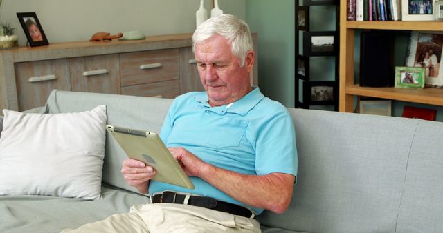 Senior man comfortably sitting on a couch while using a tablet in a modern living room. This image can be used for promoting technology use among seniors, senior lifestyles, relaxing home environments, or living room decorating ideas.