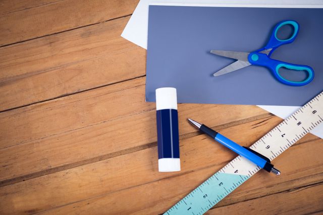 Various school supplies including scissors, a glue stick, a pen, a ruler, and papers are arranged on a wooden table. This image is ideal for use in educational materials, back-to-school promotions, crafting tutorials, or office supply advertisements.