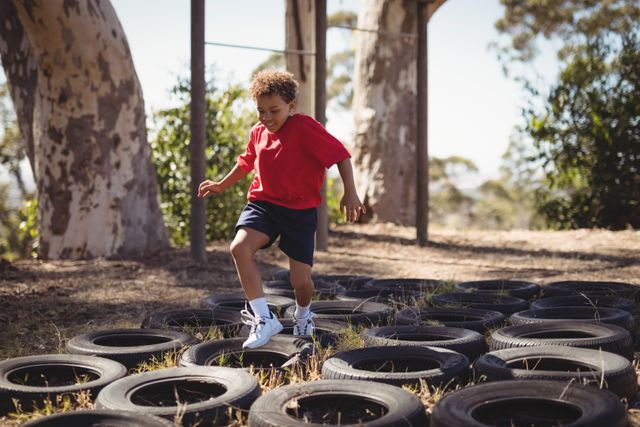 Boy running over tires in an outdoor obstacle course during a boot camp. Perfect for illustrating children's fitness activities, outdoor play, and adventure training. Ideal for use in educational materials, fitness programs, and advertisements promoting physical activity for kids.