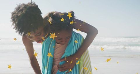 Mother wrapping child in striped towels on sunny beach with golden star confetti floating
