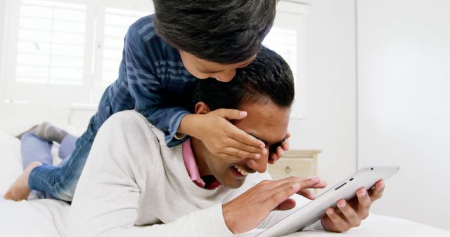 Father and son bonding moment with child playfully covering dad's eyes while he uses a tablet. Highlighting family fun with technology, this can be used for family-oriented promotions, technology in everyday life, or articles about parent-child relationships.