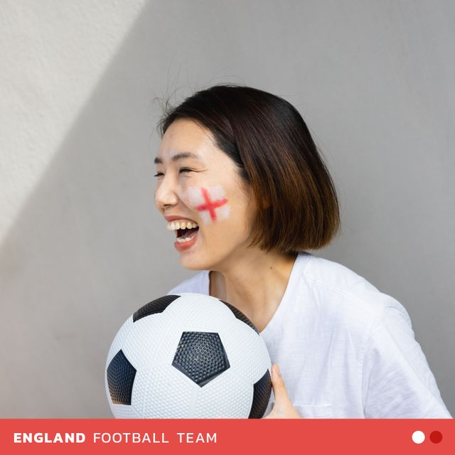 Asian female football fan showing enthusiasm by holding a soccer ball and having an England flag painted on her face. Great for use in sports team promotions, fan community events, and soccer match advertisements.