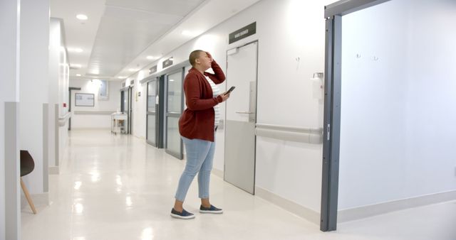 Bald woman walking down hospital corridor while looking at phone, conveying anxiety or concern. Useful for illustrating medical emergencies, healthcare environments, or personal anxiety. Great for articles about patient experiences, healthcare topics, or mobile communication.