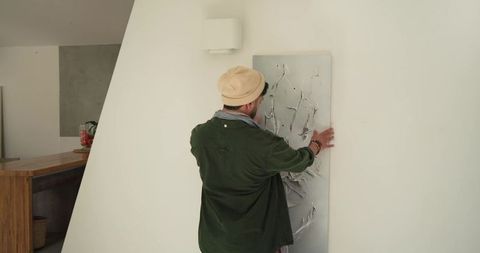 Indian man positioning tall textured abstract panel on angled white wall in modern kitchen