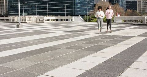 Two women walking in urban plaza wearing sportswear with phone and copy space