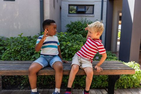 Two boys sitting on bench gesturing playfully and conversing in school courtyard amid lush greenery