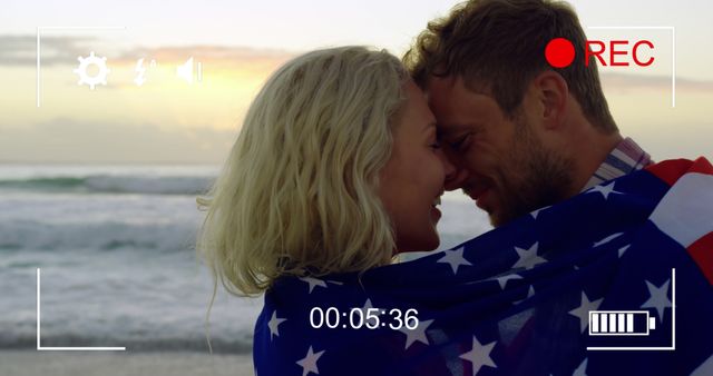 Caucasian couple embraces on the beach at sunset. Wrapped in an American flag, they share a romantic moment by the sea.
