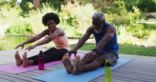 African American couple practicing yoga together on a wooden deck in a lush garden. Both are wearing comfortable workout clothes, using yoga mats, and stretching towards their toes. They appear happy and relaxed, making it ideal for promoting healthy lifestyles, exercise programs, and wellness retreats. Perfect for use in brochures, websites, and social media content related to fitness, outdoor activities, and age-inclusive health initiatives.