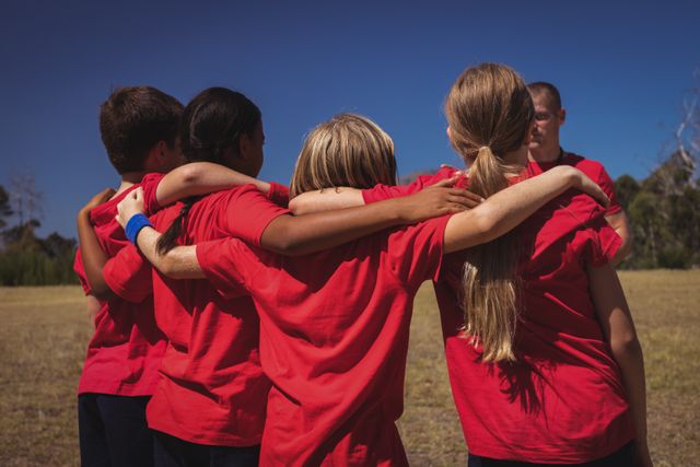 Group of children in red shirts receiving instructions from a trainer during an outdoor boot camp on a sunny day. Ideal for use in educational materials, fitness programs, team-building activities, and advertisements promoting youth sports and physical education.