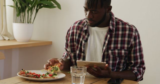 Young man casually enjoying a slice of pizza at a dining table while simultaneously using a digital tablet. This scene can be used to depict casual dining, home technology use, or everyday life. Suitable for articles or advertisements focusing on young adults, technology, or food.