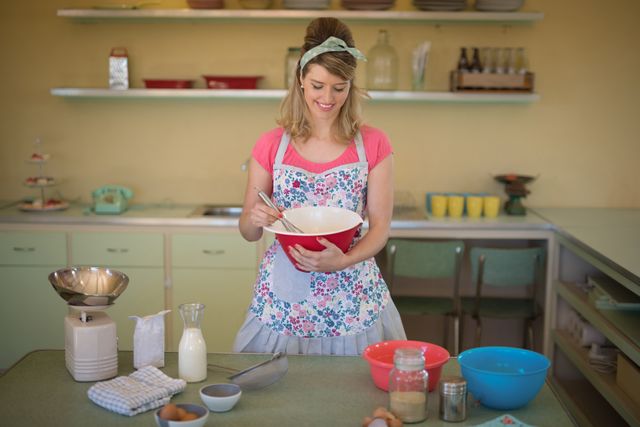 Smiling woman in a retro kitchen mixing ingredients in a bowl. She is wearing a floral apron and a headband, surrounded by baking ingredients and utensils. Ideal for themes related to vintage lifestyle, home baking, domestic life, and 1950s nostalgia.