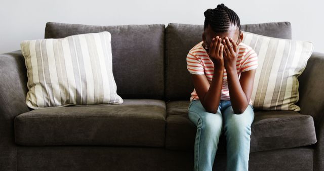 A young African American woman appears distressed, covering her face with her hands while sitting on a couch, with copy space. Her body language suggests a moment of sadness, stress, or deep contemplation.