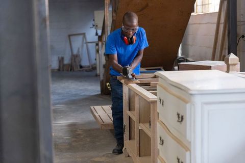 Skilled woodworker planing cabinet frame in workshop with dresser, lumber and safety gear