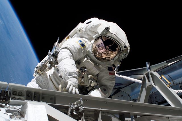 Astronaut on an extravehicular activity (EVA) or spacewalk outside the International Space Station. The astronaut is wearing a white spacesuit with Earth visible in the background. Use this for articles on space missions, NASA operations, space exploration programs, or extraterrestrial technology advancements.