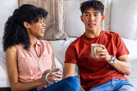 Black woman and asian man relaxing on sofa holding coffee chatting in cozy living room