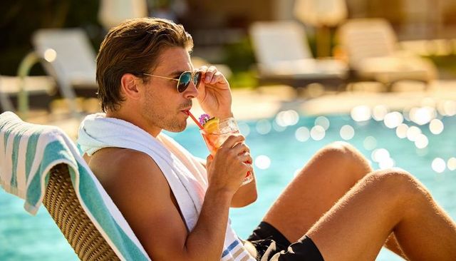 Young man relaxing at resort pool sipping cocktail and adjusting aviator sunglasses