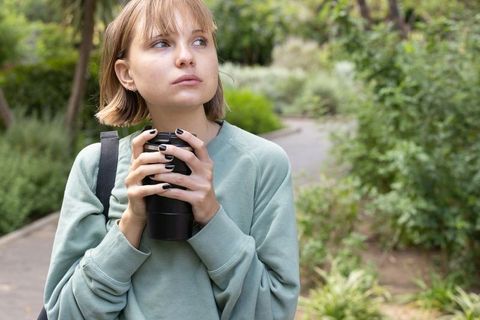 Young woman holding travel mug in park looking contemplative and cozy in green sweatshirt