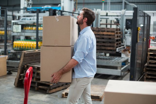 Factory worker carrying cardboard boxes in a warehouse environment. Ideal for use in articles or advertisements related to logistics, shipping, manual labor, industrial work, and warehouse operations.