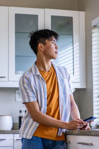 Young asian man leaning on kitchen countertop holding smartphone looking out sunlit blinds
