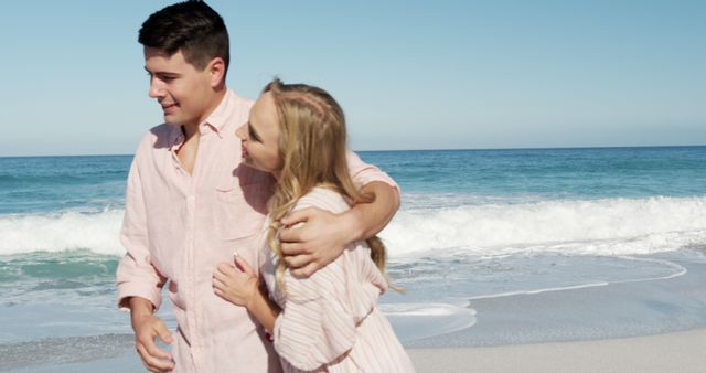 Young couple walking together on sandy beach by ocean waves, enjoying good weather, hugging each other in affectionate mood. Ideal for travel advertisements, romantic getaways, summer promotions, and lifestyle blogs.