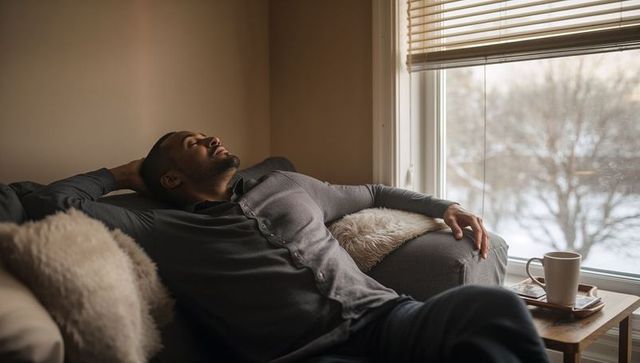 Reclining man relaxing on cozy gray sofa by snowy window with coffee and fuzzy pillows