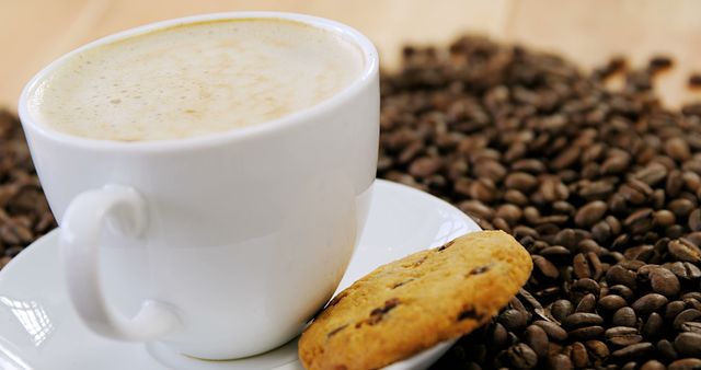 White coffee cup on saucer with frothy coffee next to a chocolate chip cookie, surrounded by coffee beans. Ideal for illustrating coffee shop menus, breakfast promotions, cafe advertisements, or food and beverage blogs.