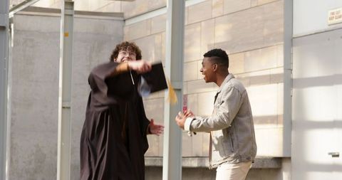 Graduate tossing mortarboard while friend cheering in sunlit campus courtyard
