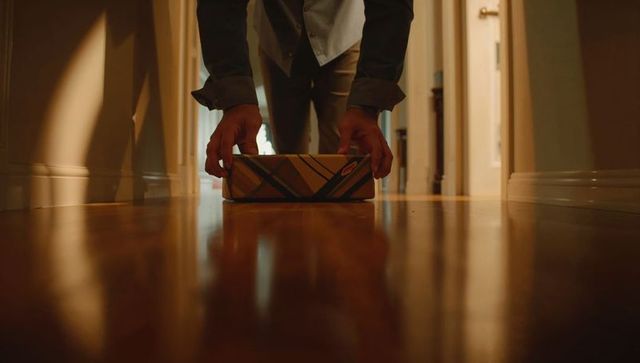 Man examining striped shoebox on polished hardwood hallway floor in warm amber light