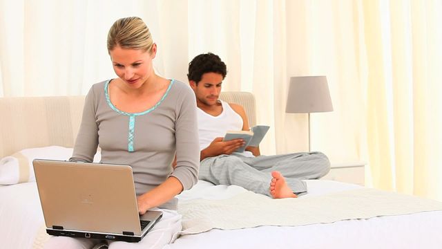 Young couple relaxing in a cozy bedroom; woman focused on using a laptop while the man reads a book. Ideal for depicting modern lifestyle, leisure time activities, comfortable living spaces, and work-life balance.