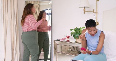 African american friends applying makeup and styling hair in bright bedroom mirror