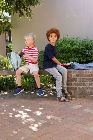 Multicultural school boys sitting on bench with backpacks and tablet in shaded courtyard