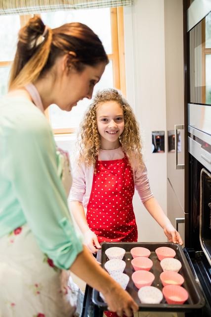 Mother and daughter are baking cupcakes together in a home kitchen. The daughter is holding a tray with cupcake liners, and both are wearing aprons. This image is perfect for illustrating family bonding, home cooking, and domestic life. It can be used in articles, advertisements, or social media posts related to family activities, baking recipes, or parenting tips.