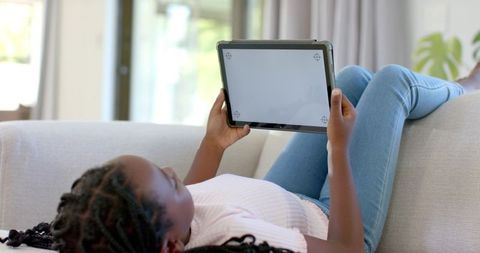 African american girl lying on sofa using tablet for learning and entertainment at home