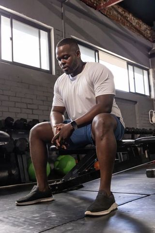 African american man resting on bench checking smartwatch in brick-walled industrial gym with weight
