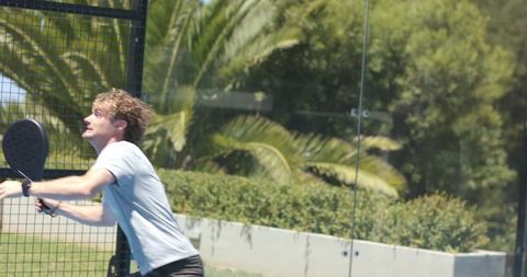 Male playing padel reaching overhead shot on sunlit outdoor glass court with wristwatch