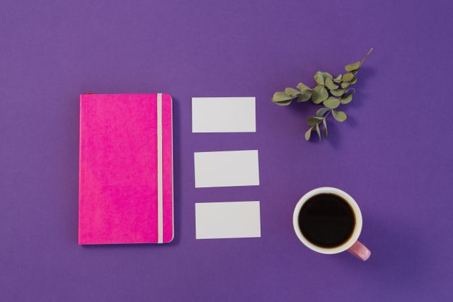 Overhead view of a workspace featuring a pink notebook, three blank business cards, a cup of black coffee, and a sprig of dry leaves on a purple background. Ideal for use in articles or advertisements related to office organization, productivity, or creative workspaces.