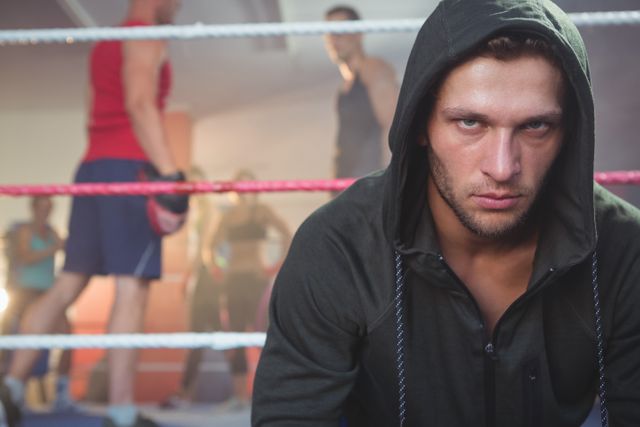 Young male boxer in a hoodie sitting on a boxing ring, looking determined and focused. Background shows other athletes training, creating a dynamic and energetic atmosphere. Ideal for use in fitness, sports, and motivational content, as well as advertisements for gyms, boxing clubs, and athletic wear.