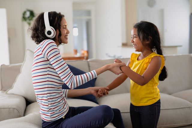 Happy hispanic mother wearing headphones, playing with daughter in living room. family spending time together at home.