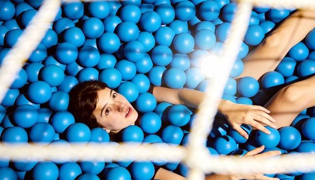 Young woman reclining in sea of blue play balls reaching through white net with lens flare
