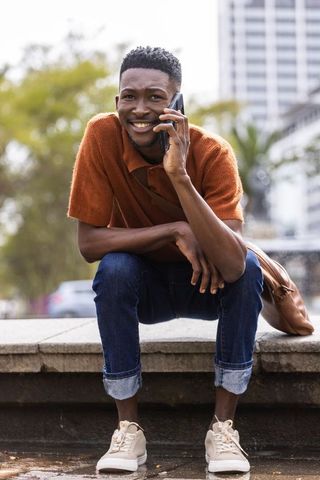 Smiling man talking on phone while crouching by urban plaza fountain with backpack