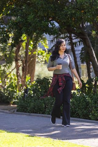 Woman walking in sunlit park holding takeaway coffee, casual outfit with plaid shirt tied