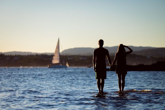 This image captures a romantic moment of a couple standing by a serene lake during sunset, holding hands and gazing at a sailboat in the distance. Ideal for travel brochures, romantic getaway promotions, relationship blogs, and lifestyle magazines focused on outdoor leisure and relaxation.
