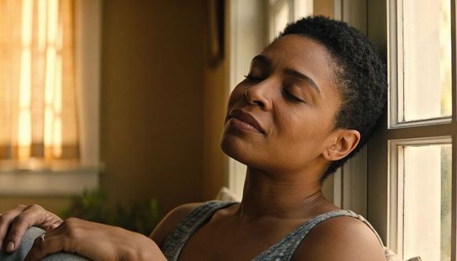 Woman leaning back by sunlit window, closing eyes in quiet contemplation