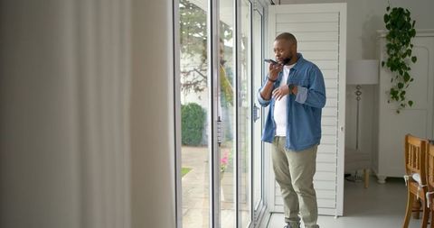 African american man talking on smartphone while standing by sliding glass doors at home