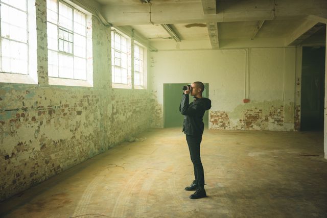 Young man taking photos in an empty, abandoned warehouse with an SLR camera. He is wearing a black jacket and standing in a spacious, industrial setting with brick walls and large windows. Ideal for themes of urban exploration, creativity, solitude, and industrial photography.