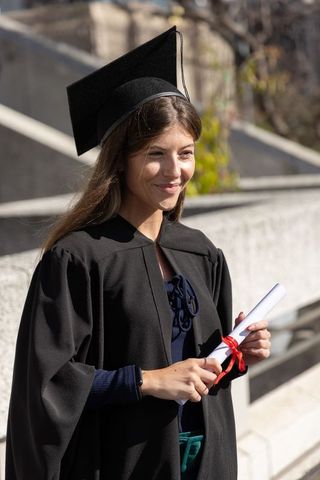 Smiling female graduate wearing cap and gown holding diploma on campus stone steps